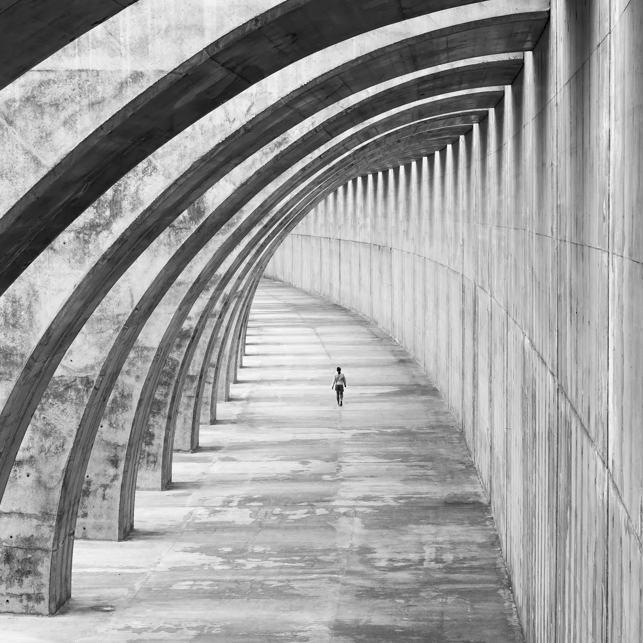 Arches de béton formant un couloir courbe, un promeneur seul au loin